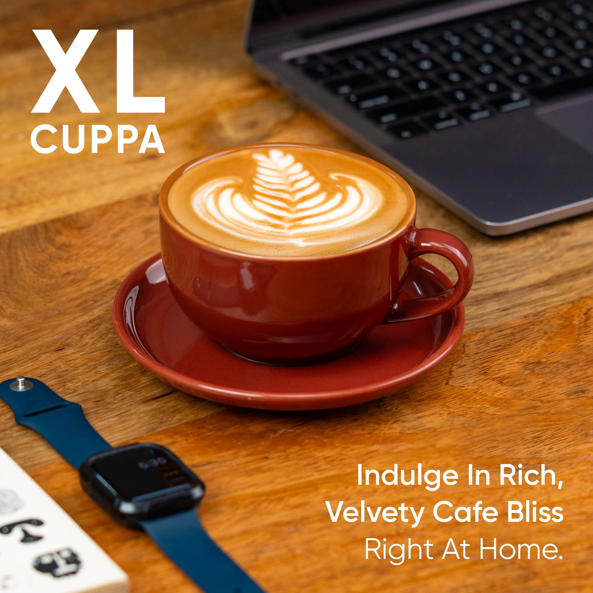 Red cup of cappuccino with latte art on a wooden table, accompanied by a laptop and smartwatch.