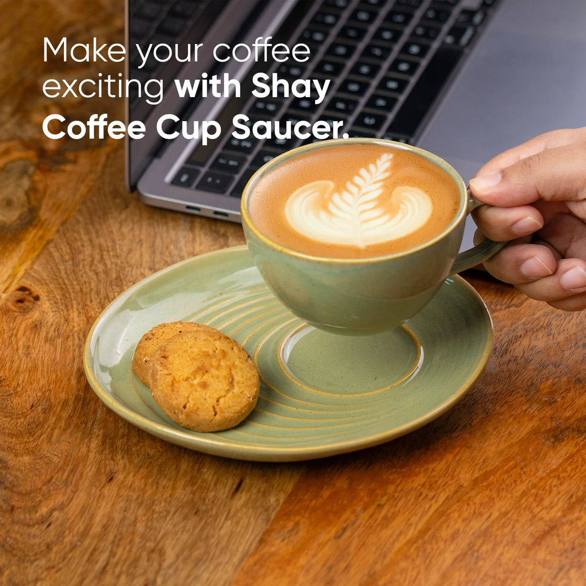 Cup of coffee with a leaf design on a saucer, held by a hand, with a laptop in the background.
