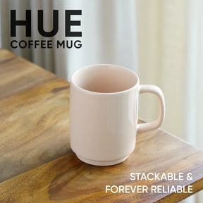 White coffee mug on a wooden surface with 'HUE' branding and text about stackability and reliability.
