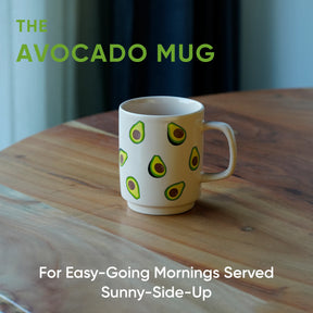 White mug with avocado design on a wooden table, text 'The Avocado Mug' and 'For Easy-Going Mornings Served Sunny-Side-Up'.