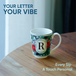 Decorative mug with floral design and letter 'R' on a wooden surface, with text 'Your Letter Your Vibe' and 'Every Sip A Touch Personal'.