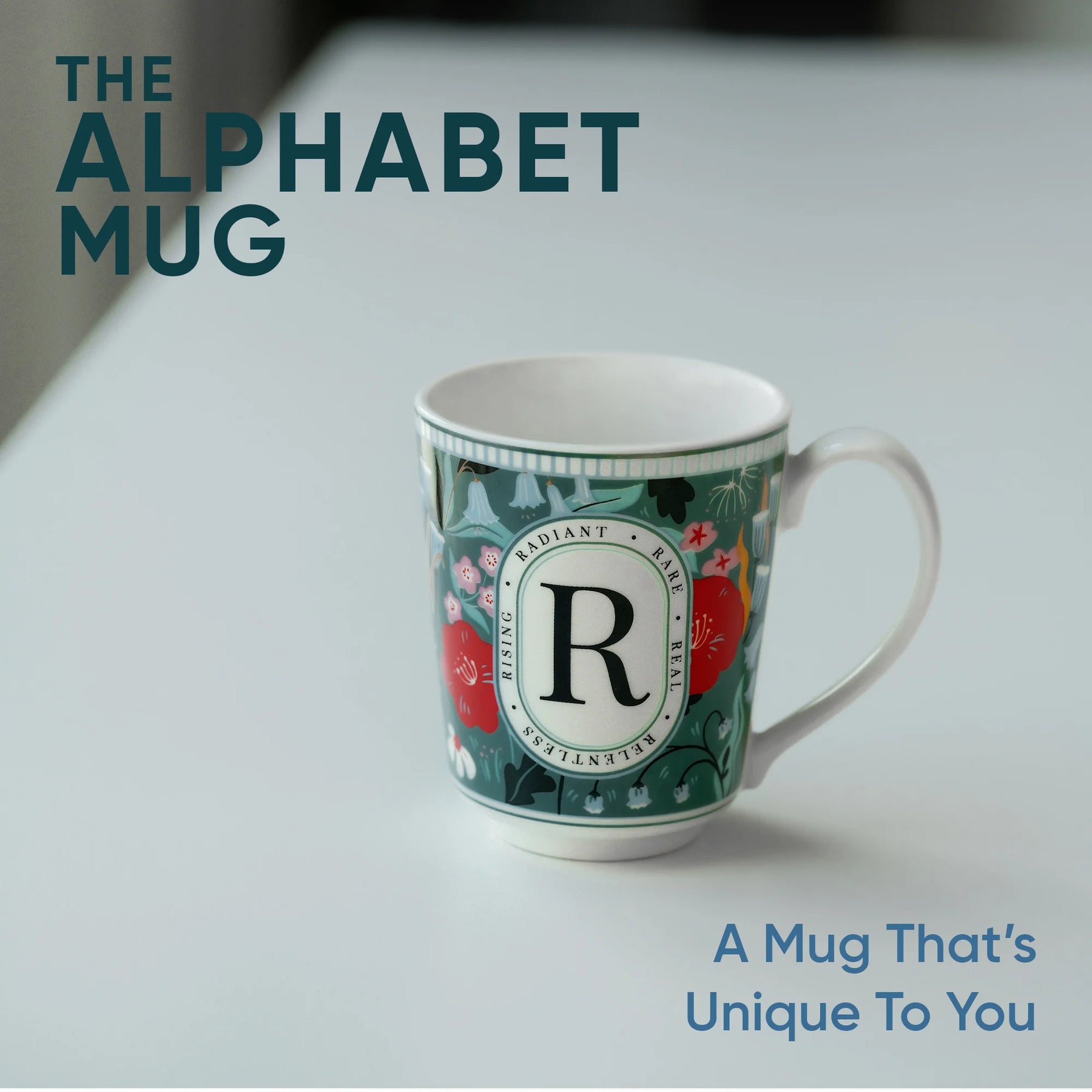 Mug with floral design and letter 'R' on a white surface, text 'The Alphabet Mug' and 'A Mug That's Unique To You' displayed.