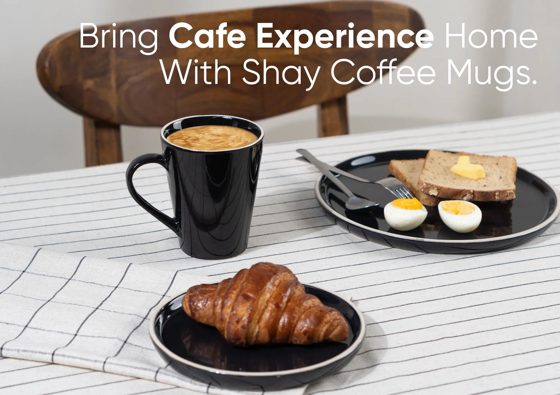 Black coffee mug with coffee, a croissant, and a plate with eggs and toast on a table.